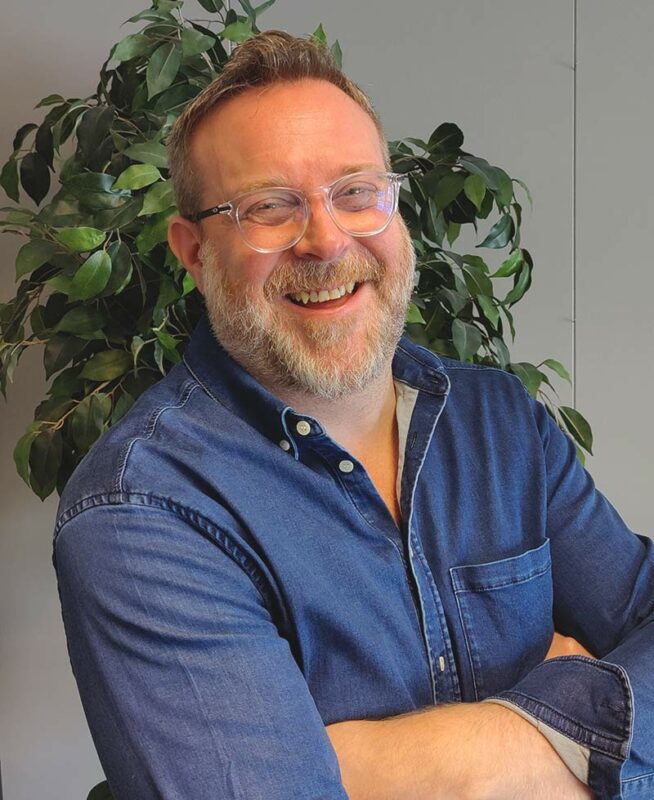 a man with a beard and glasses sitting in front of a plant.